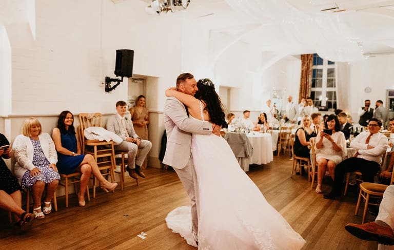 Bride and groom having first dance in village hall surrounded by guests