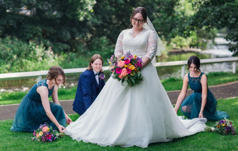 Bride stood on lawn smiling holding flowers with bridesmaids kneeling tending to hem of dress