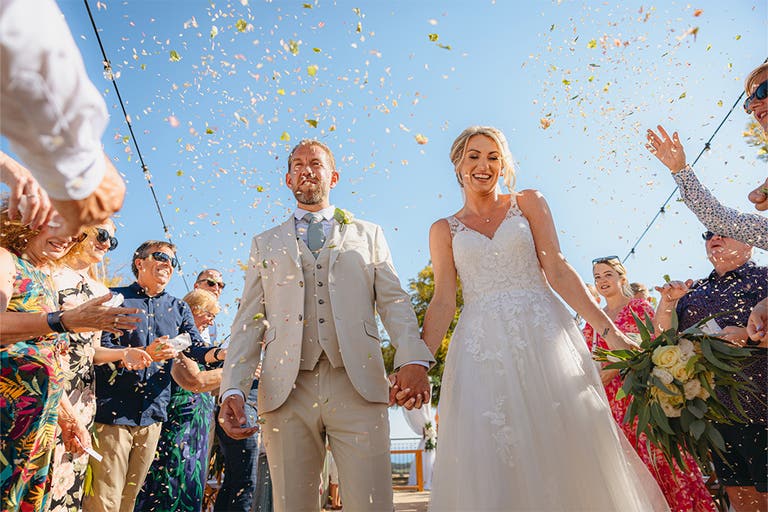 Katie and James holding hands while confetti is being thrown by their wedding guests