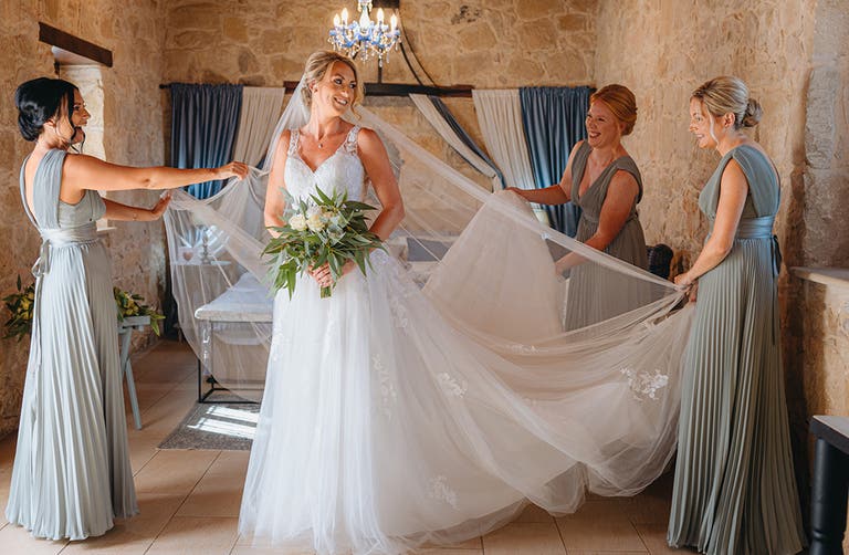 Katie standing in the bridal suite in her wedding dress with the bridesmaids fanning out her train and veil