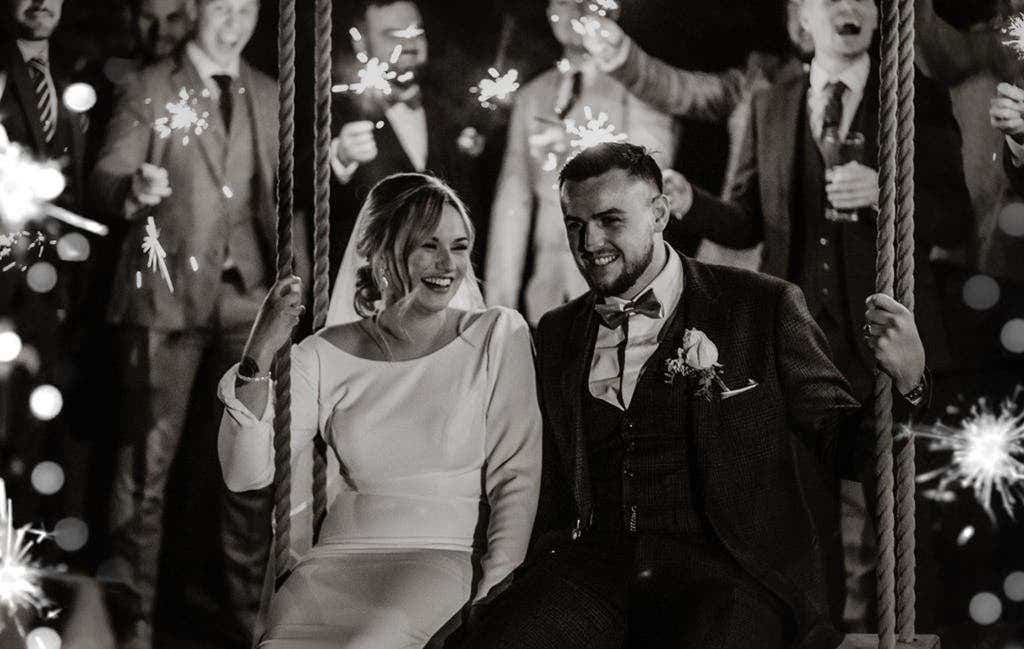 Brid and groom sat smiling on rope swing surrounded by guests holding sparklers 