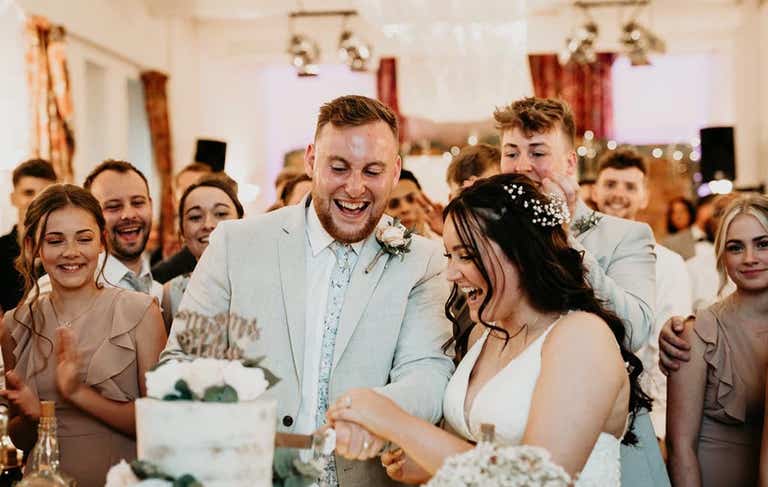Bride and groom laughing cutting cake while guests look on cheering