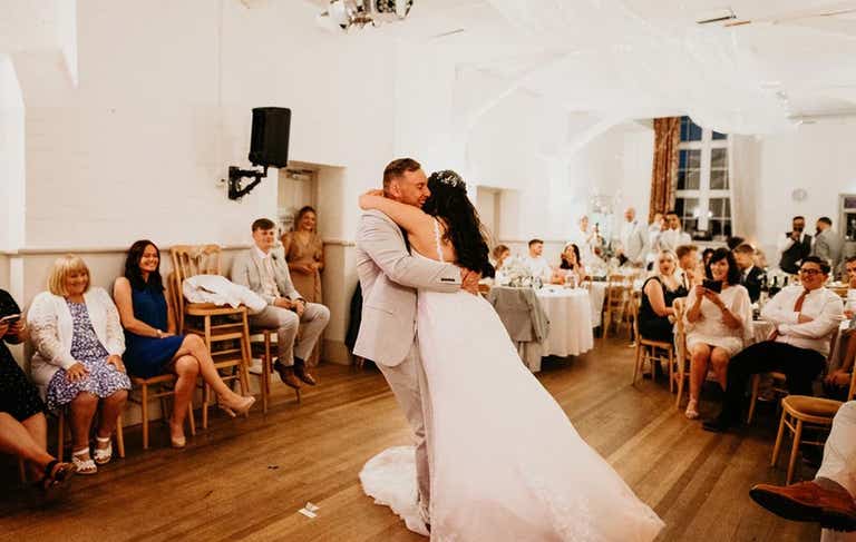 Bride and groom having first dance in village hall surrounded by guests
