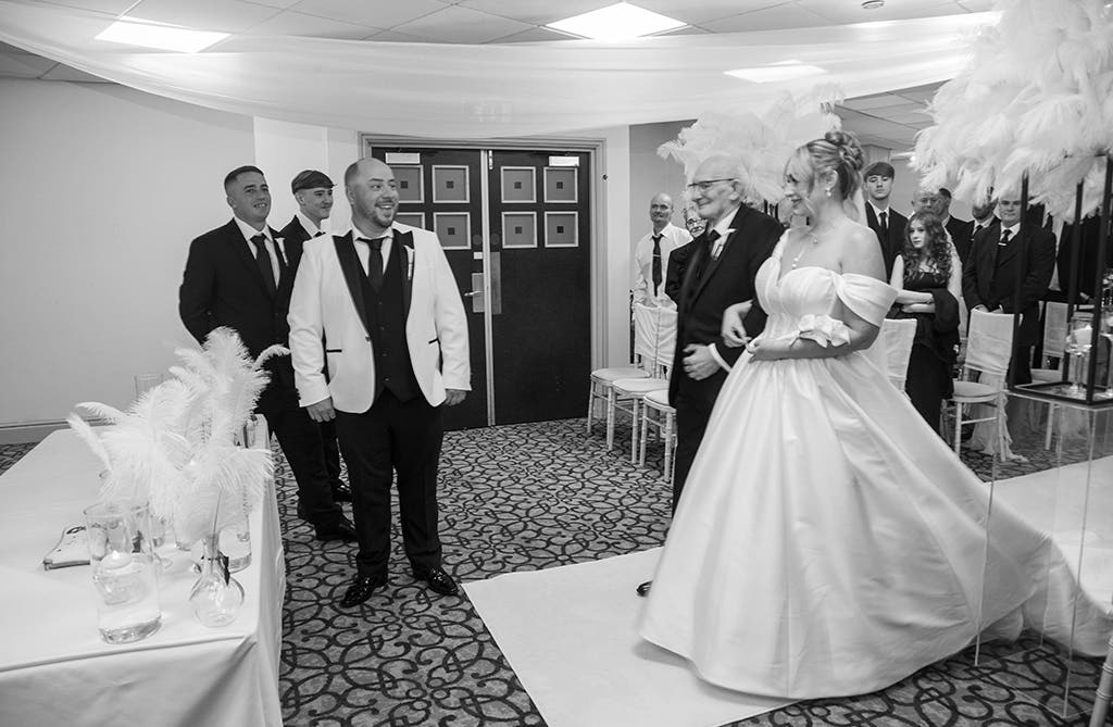 Black and white image of bride Rachel walking down the aisle with Andrew smiling during their hotel wedding ceremony