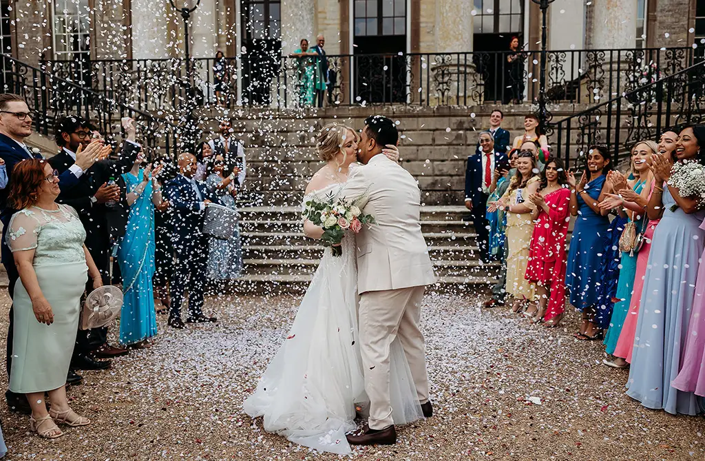 Fran and Ronit in Western wedding attire outside Ragley Hall with guests throwing confetti