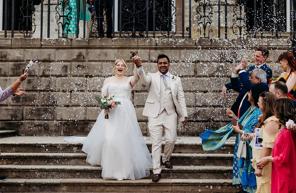 Fran and Ronit walking down the steps of Ragley Hall in Western wedding attire with confetti